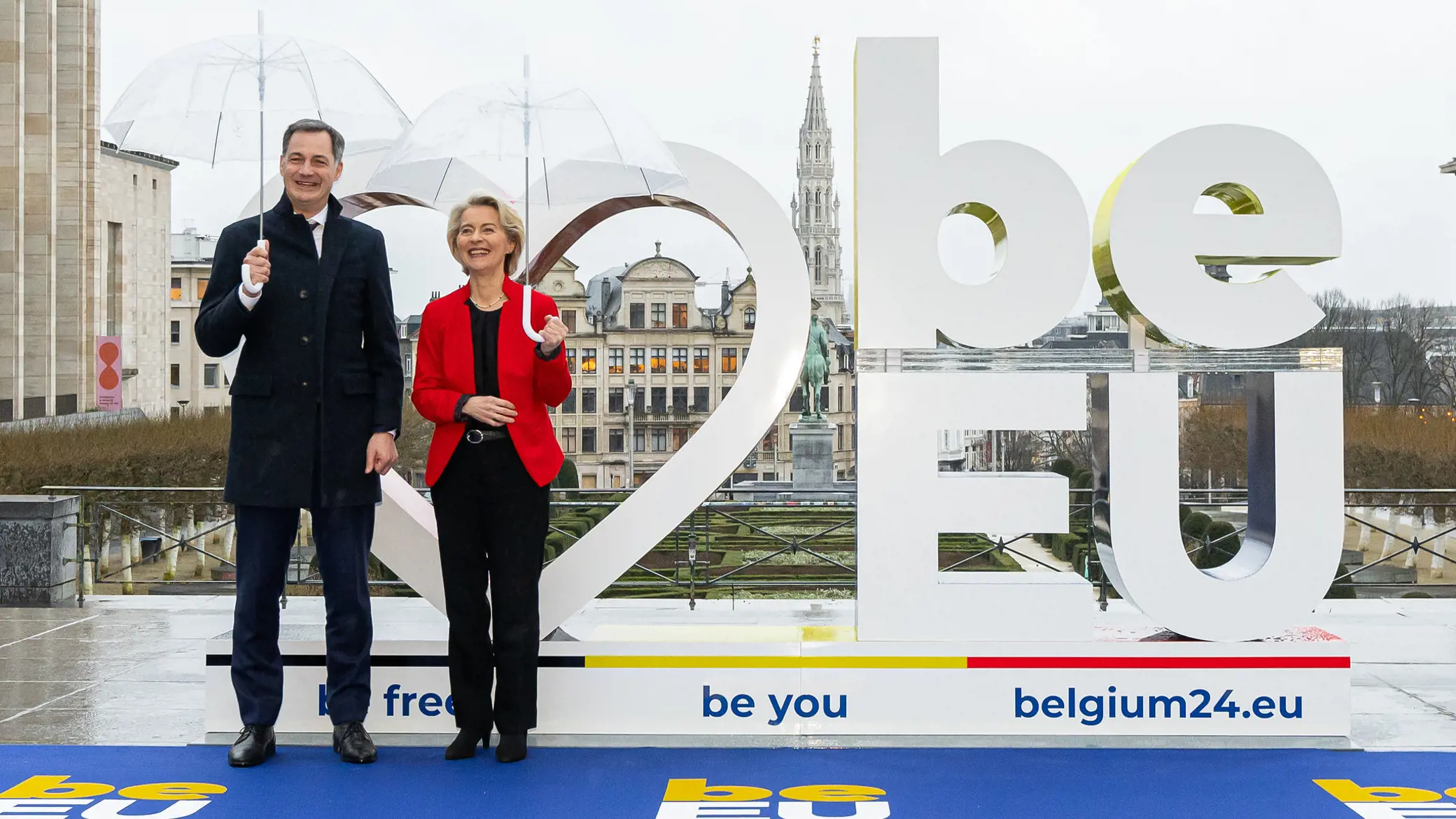 Photo of Ursula von der Leyen and Alexander De Croo stading with umbrellas welcoming the Belgian Presidency launch by a BE EU 2024 artistic logo sculpture in a grey and rainy Brussls. Photo credit @ Belgian Presidency.